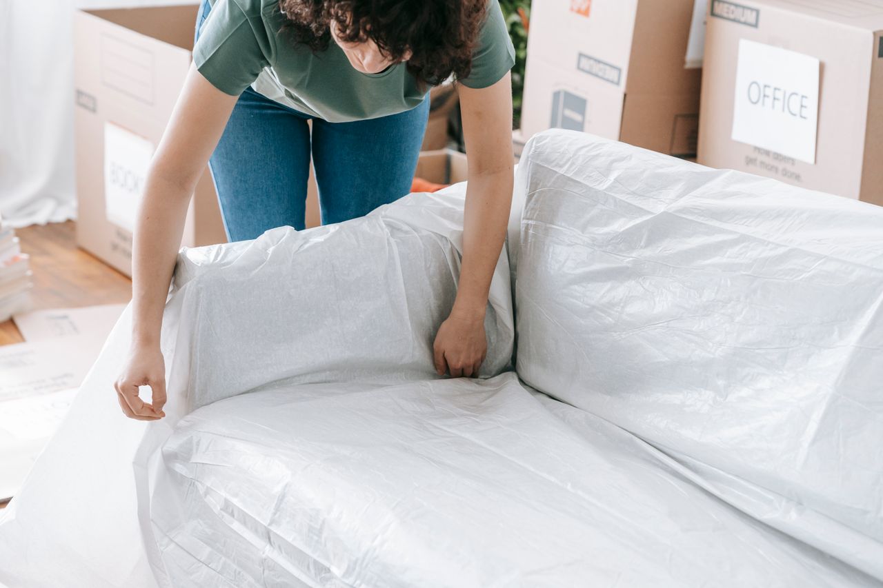 why-choose-us A woman carefully wraps a sofa in protective material while packing boxes in a bright room.